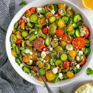 Tomato cucumber Mediterranean salad in a white bowl with a serving spoon topped with feta cheese and olives.