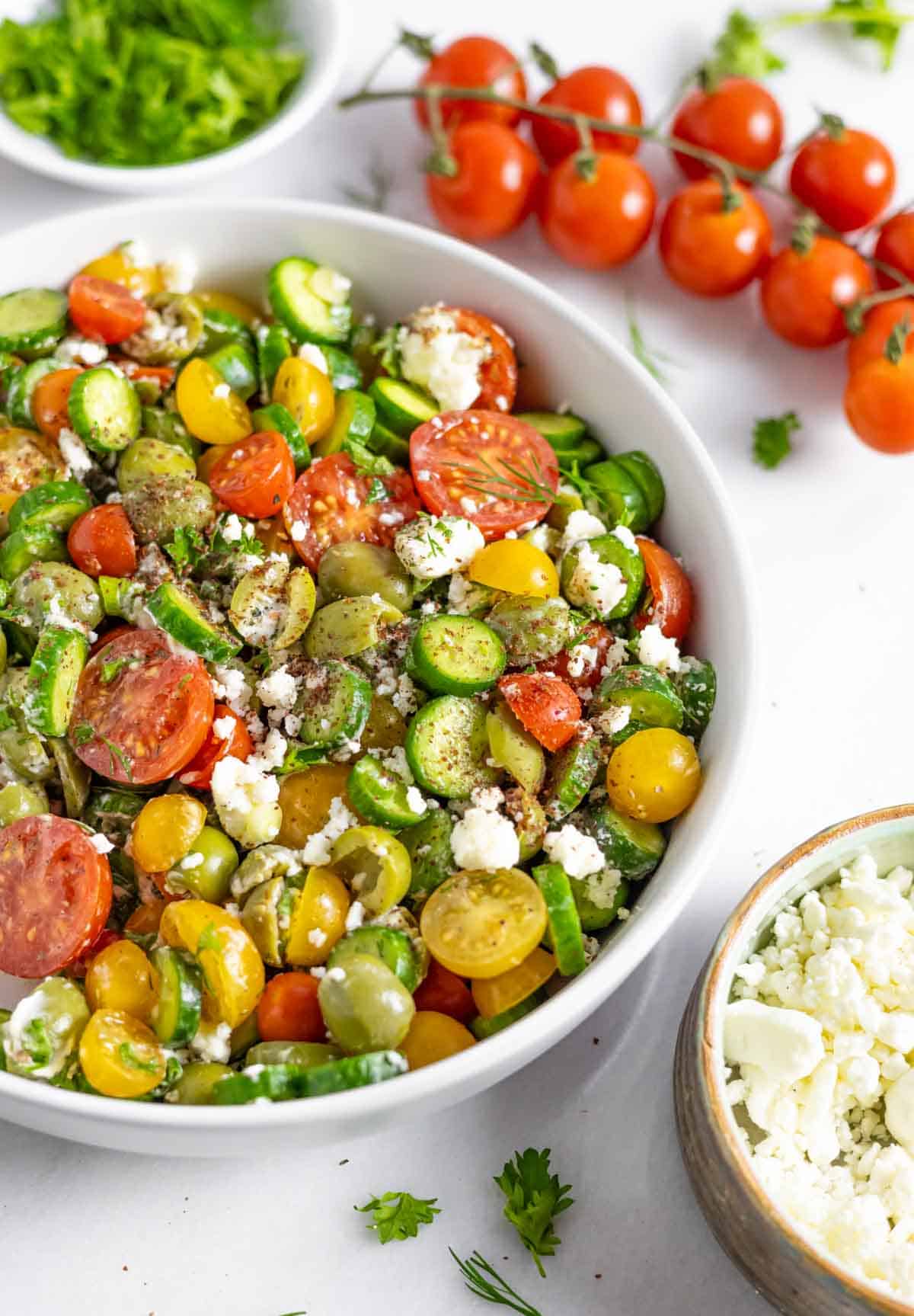 Bowl of Mediterranean cucumber and tomato salad next to cherry tomatoes on the vine and a small bowl of feta cheese.