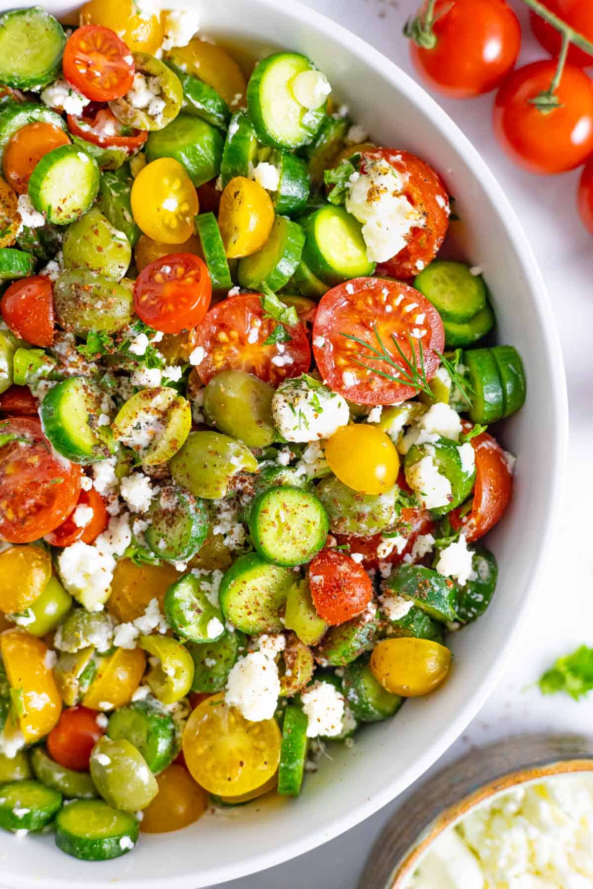 Close up of half a white bowl of tossed greek salad with cherry tomatoes on the vine next to the bowl.