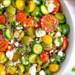 Close up of half a white bowl of tossed greek salad with cherry tomatoes on the vine next to the bowl.