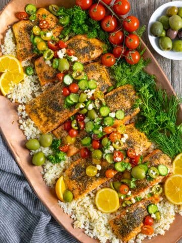 Seven pieces of Mediterranean seasoned steelhead trout on a bed of cauliflower rice on a copper tray garnished with tomato cucumber salad.