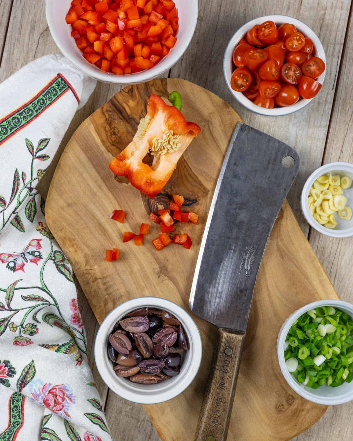 Small bowls of chopped ingredients, on and next to a wood board with a cleaver, and a red pepper partially chopped.