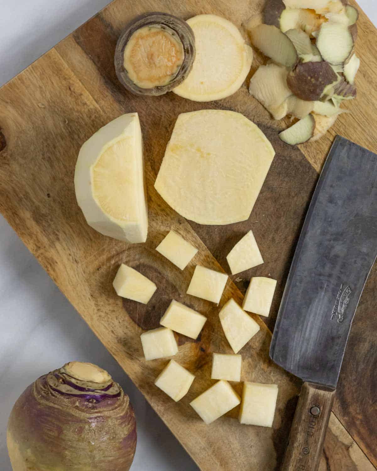 Peeled rutabaga cut in half on a wood board and partly chopped into 1 inch chunks next to a cleaver.