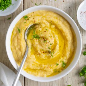 White bowl of rutabaga puree with a spoon, topped with melted butter and sprinkled with herbs.