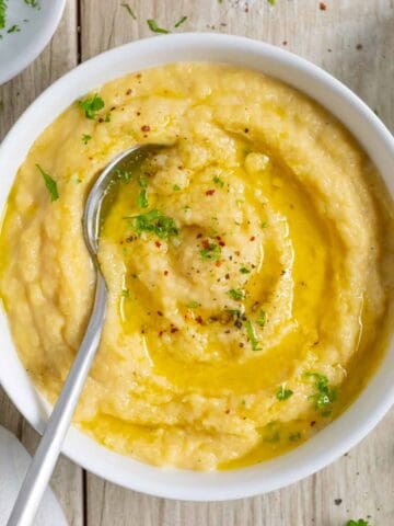 White bowl of rutabaga puree with a spoon, topped with melted butter and sprinkled with herbs.