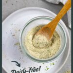Glass bowl of homemade ranch mix with a small wood spoon in it, sitting on a white plate sprinkled with herbs.