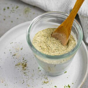 Glass bowl of homemade dairy free ranch seasoning with a small wood spoon in it, sitting on a white plate sprinkled with herbs.
