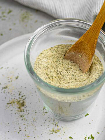 Glass bowl of homemade dairy free ranch seasoning with a small wood spoon in it, sitting on a white plate sprinkled with herbs.