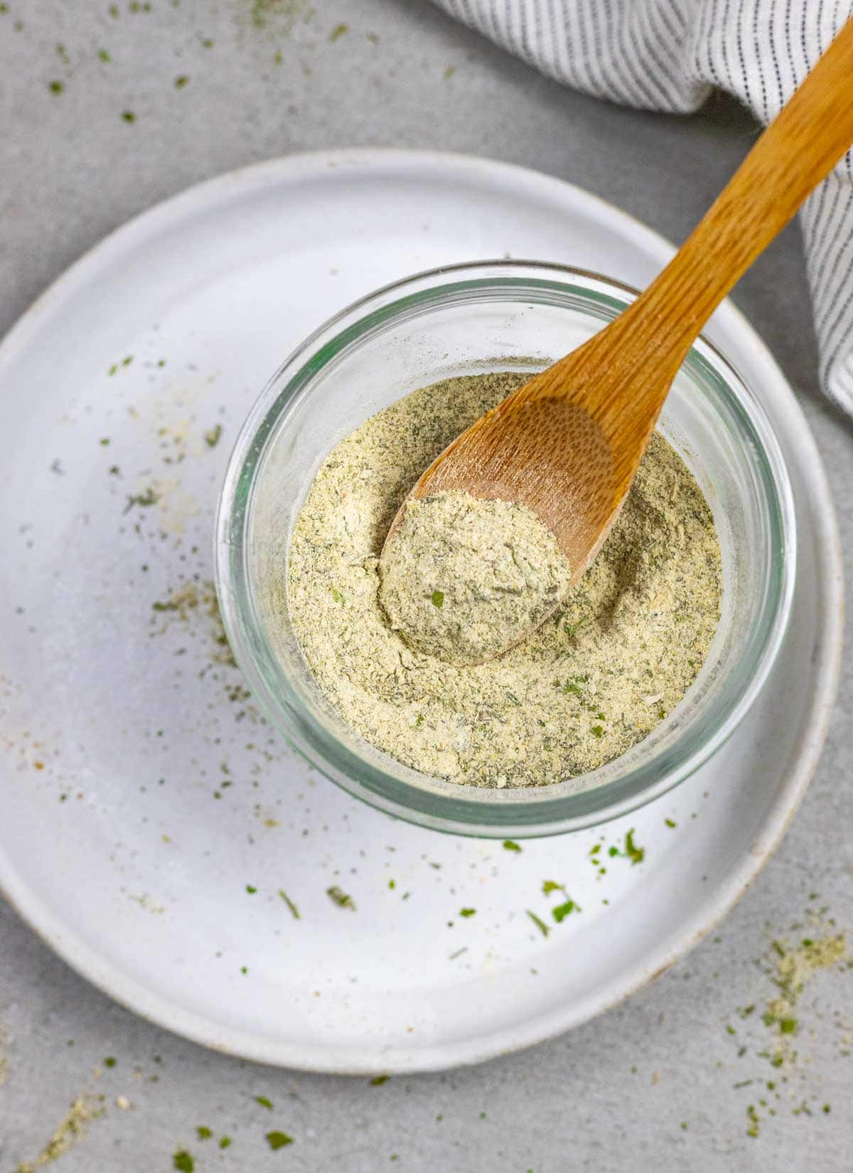 Glass bowl of homemade ranch mix with a small wood spoon in it, sitting on a white plate sprinkled with herbs.
