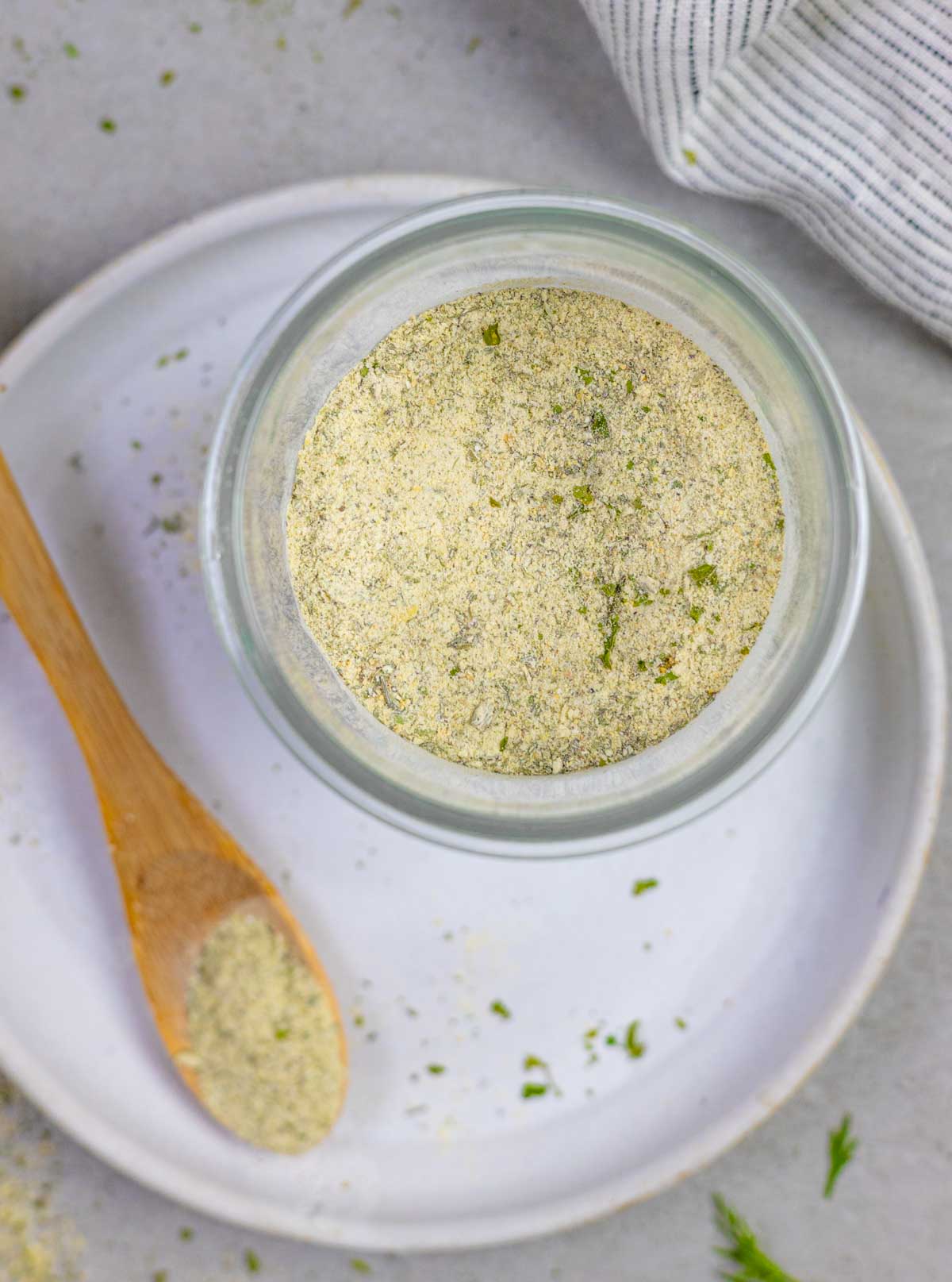 Glass bowl of homemade ranch mix on a white plate sprinkled with herbs and a small wood spoon with seasoning on it.