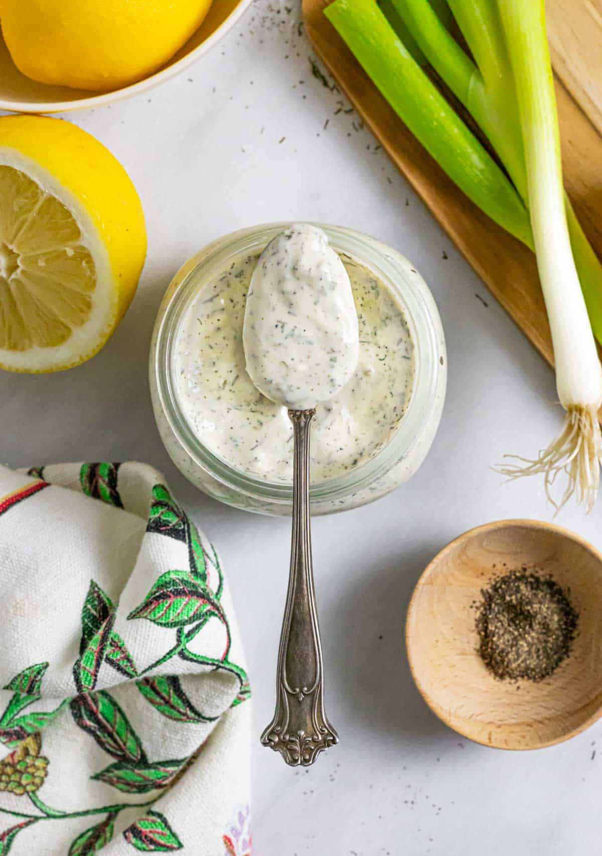 Spoon of ranch dressing balanced over the mouth of a full jar, on a board with spices in small bowls.